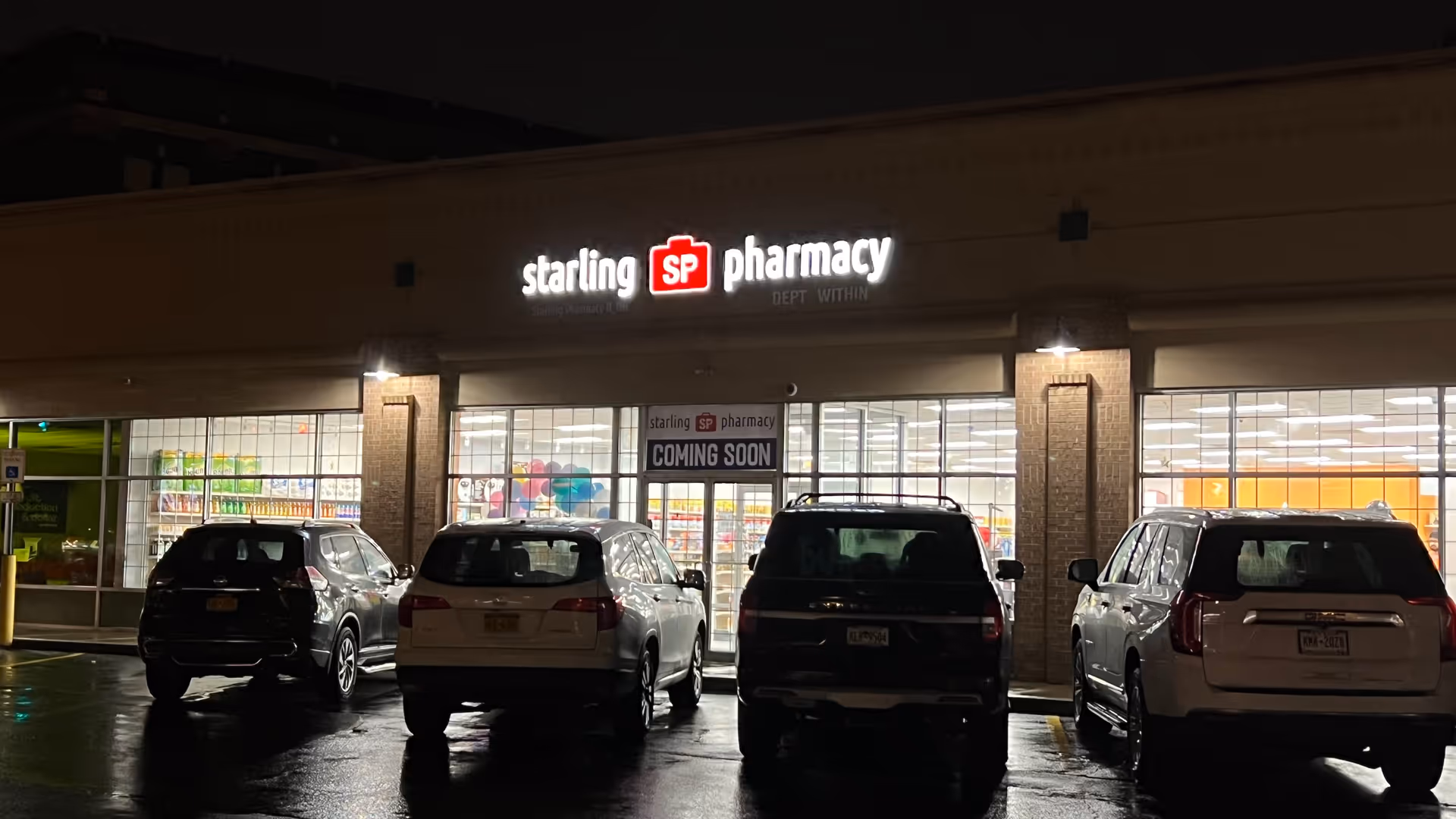Night exterior view of a storefront with Starling SP Pharmacy sign and parked cars