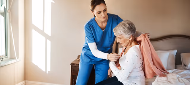 Caregiver in blue scrubs assists elderly woman in a cozy bedroom.