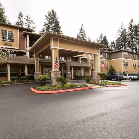 Exterior view of a senior living facility with a covered entrance supported by stone pillars, two American flags displayed, landscaped greenery, and parked vehicles in the background under an overcast sky.