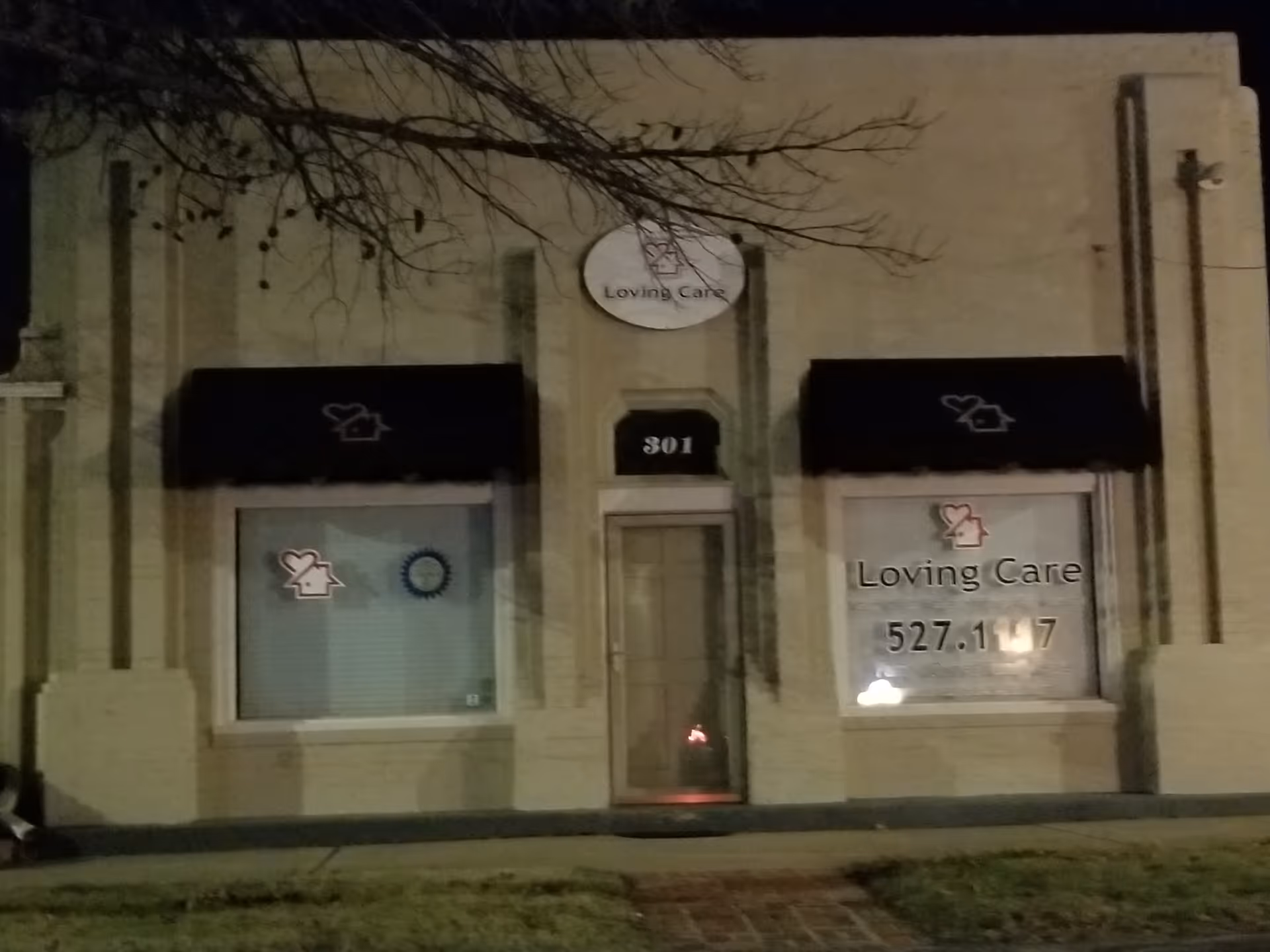 Nighttime exterior of a beige building with two storefront windows, door, and Loving Care signage.