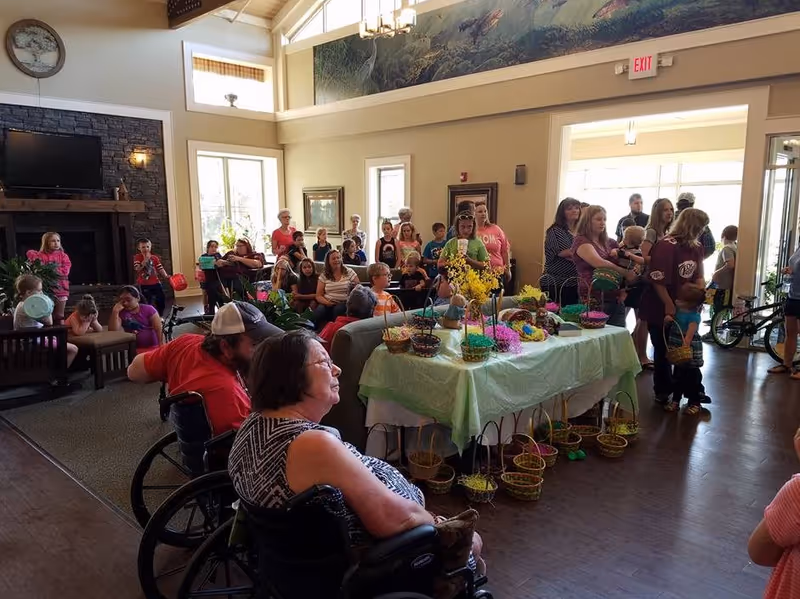 A large group of people, including children and elderly individuals, gathered in a spacious, well-lit room with high ceilings and large windows. Several people are seated, including two individuals in wheelchairs in the foreground. A table covered with a green cloth is decorated with colorful baskets and flowers. The room has a cozy fireplace with a TV above it and framed artwork on the walls.