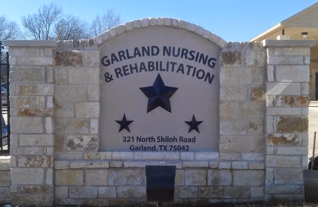 Stone monument sign for Garland Nursing & Rehabilitation Center with three stars and the address 321 North Shiloh Road, Garland, TX 75042, set outdoors with trees and part of a building visible in the background.