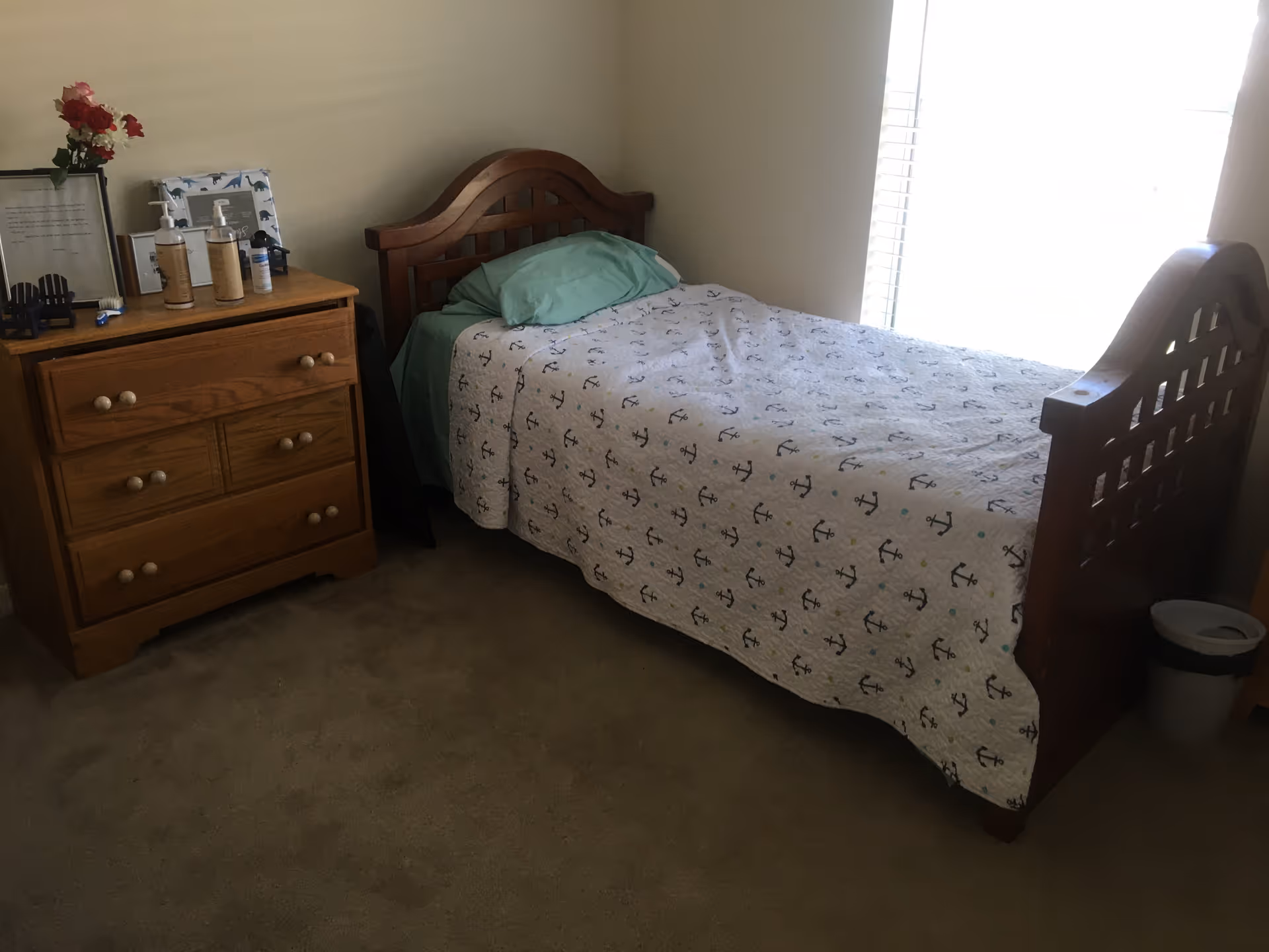 Bedroom with wooden bed, dresser, anchor-patterned quilt, teal pillows, and window light.
