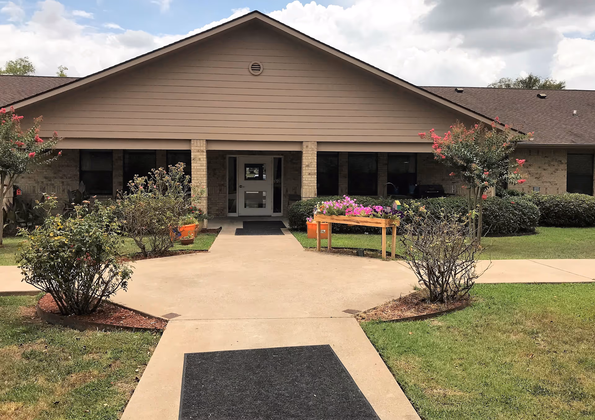 Front entrance of a single-story brick nursing facility with a paved walkway, planted flower beds and a bench with potted flowers.