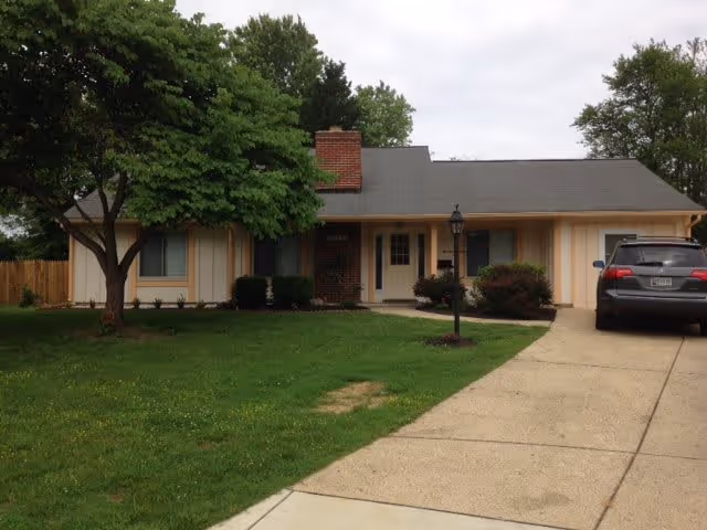 Single-story ranch-style house with a front lawn, driveway, lamp post and a parked car under an overcast sky.