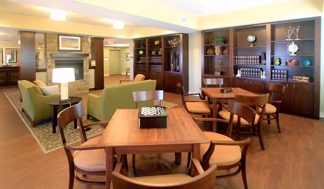 A cozy common area in a senior living facility featuring wooden tables and chairs arranged for games or socializing. There are two green armchairs and a small side table with a lamp near a stone fireplace. Built-in wooden bookshelves with decorative items and books line the back wall. The room has warm lighting and a wooden floor.