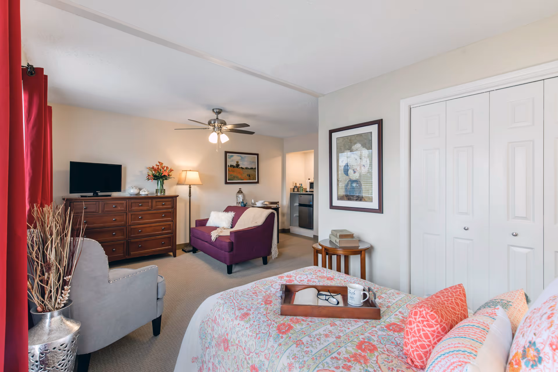 Bedroom in a senior living suite with floral bedspread, bed, dresser, purple seating, and closet doors.