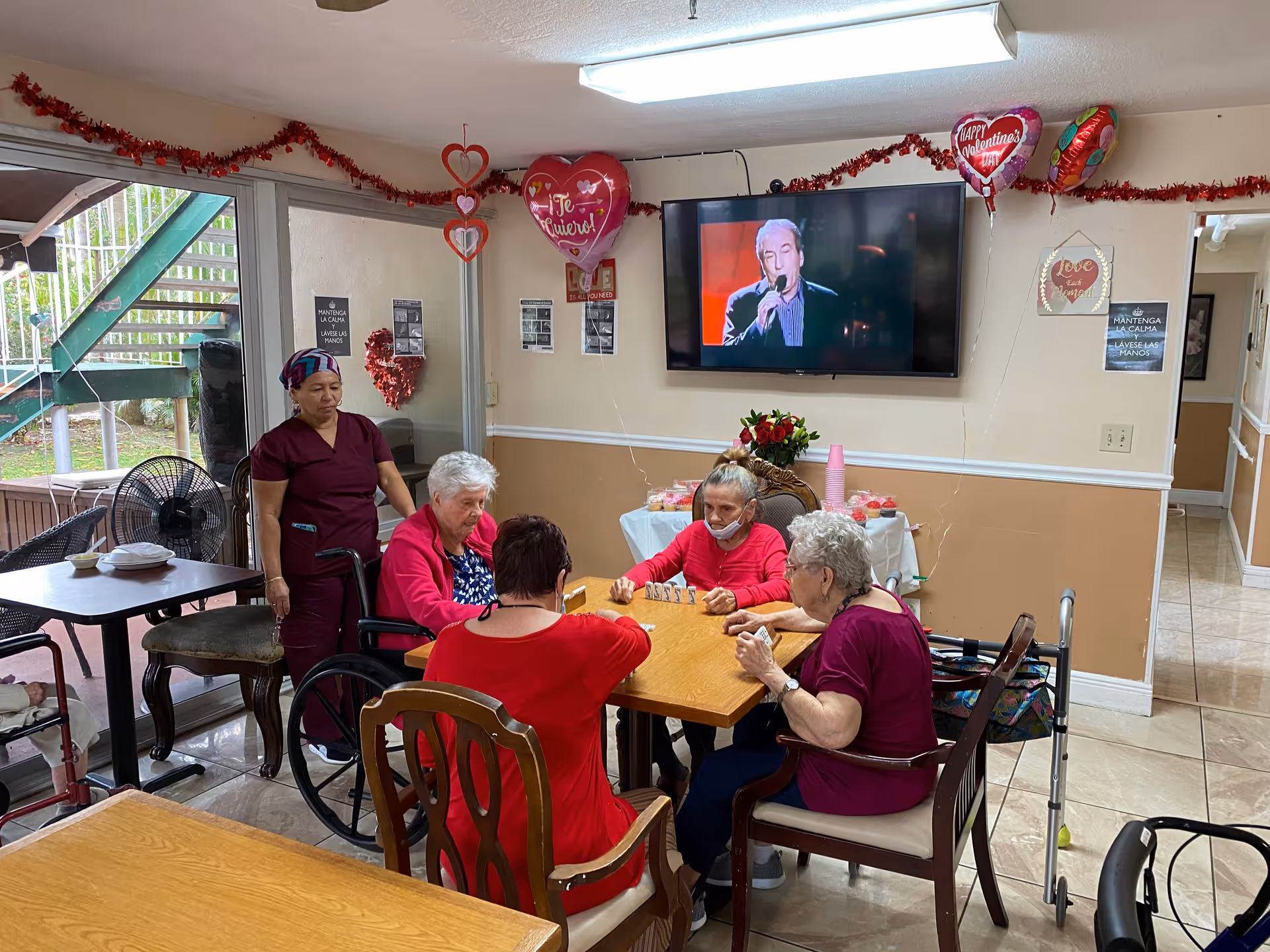 A group of elderly women sitting around a wooden table playing a game in a decorated room. A caregiver stands nearby. The room has Valentine's Day decorations including heart-shaped balloons and garlands. A flat-screen TV mounted on the wall shows a man singing. There is a table with pink cups and flowers in the background, and a glass door leading outside.