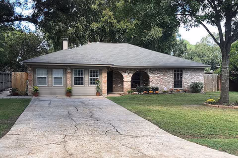 Single-story brick ranch-style building with a long cracked concrete driveway, front lawn, potted plants, and trees.