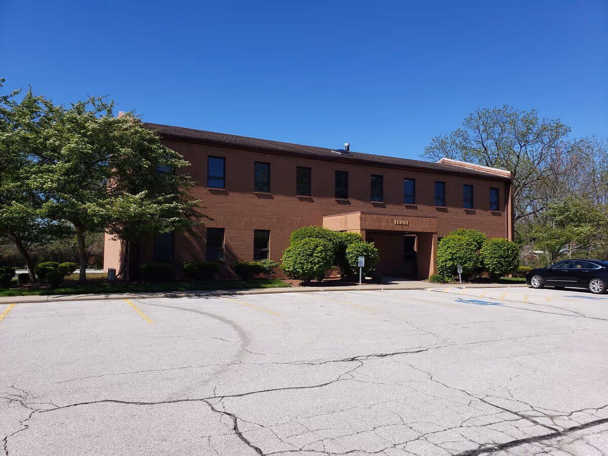 Brick two-story facility entrance with manicured shrubs, trees, and a parking lot.