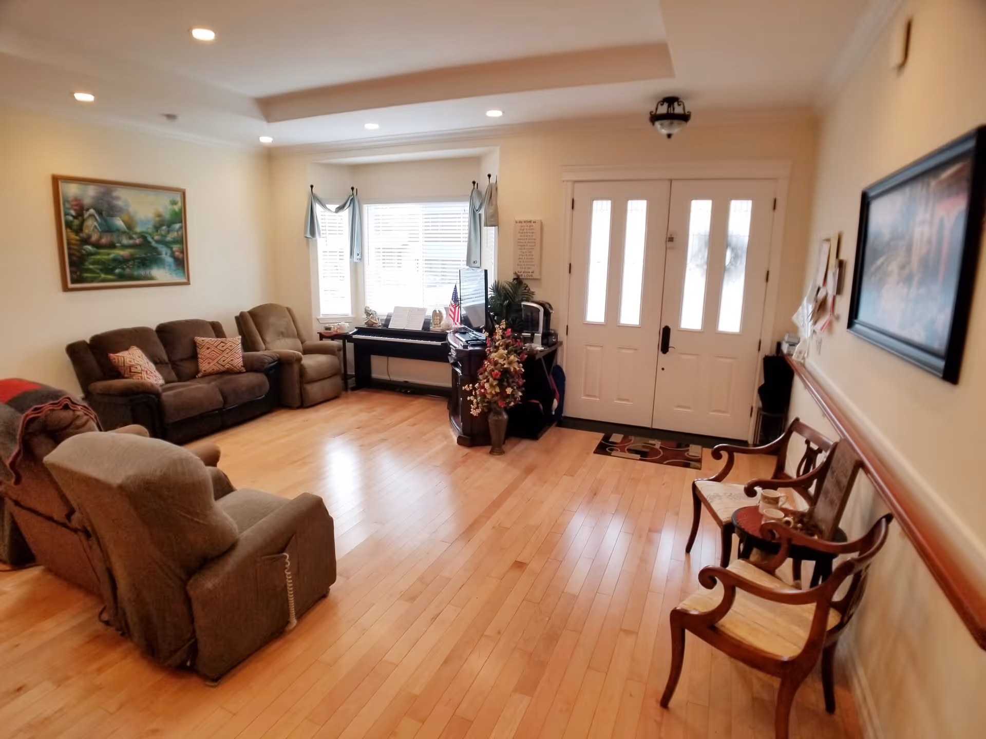 A cozy senior living room with wooden flooring, a brown sofa set with cushions, two armchairs, a small table with a cup, and a piano near a window with white blinds. The room has a white front door with glass panels, wall paintings, and a vase with flowers near the piano.