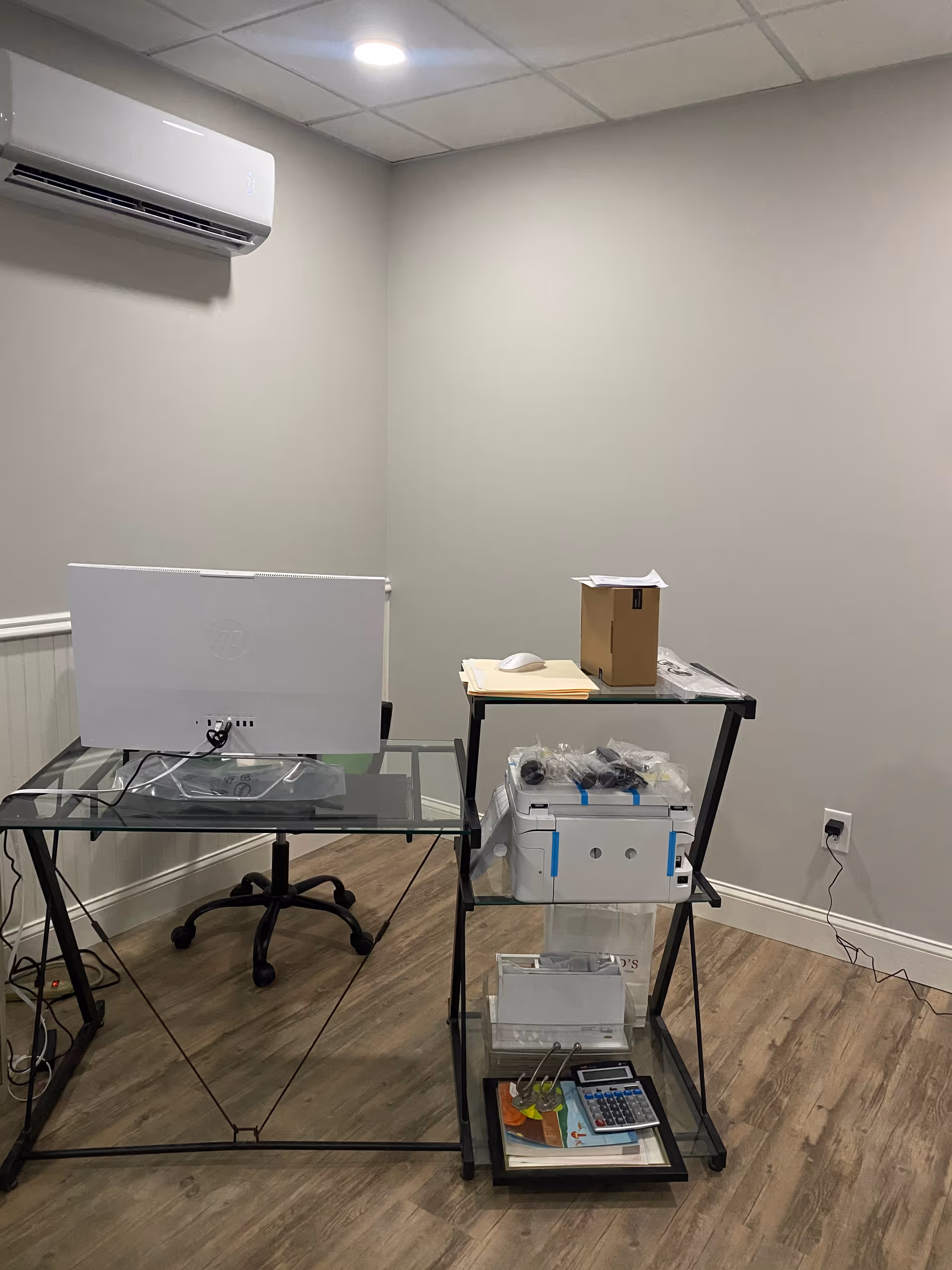 A small office setup with glass desk, computer monitor, and a rolling cart