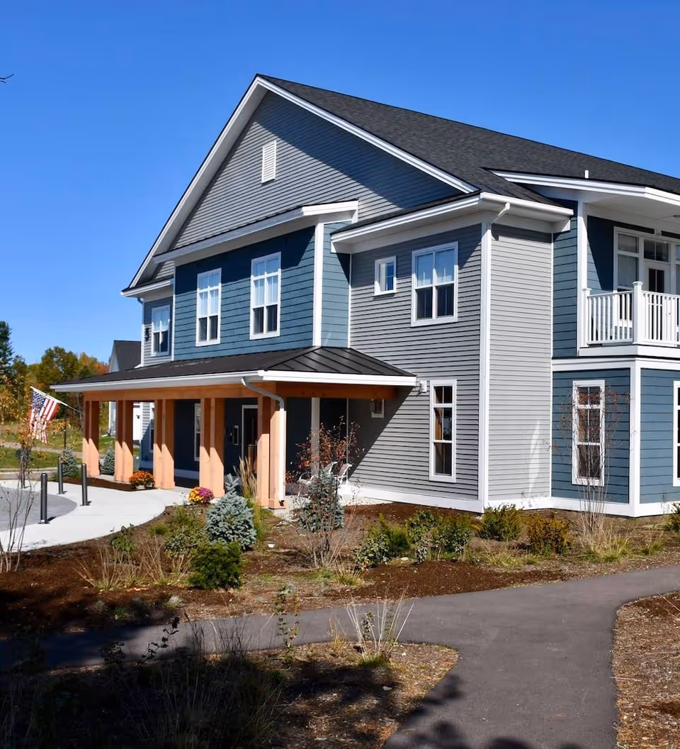 Two-story blue-gray building with a covered entrance, balcony, American flag, and landscaped paths.