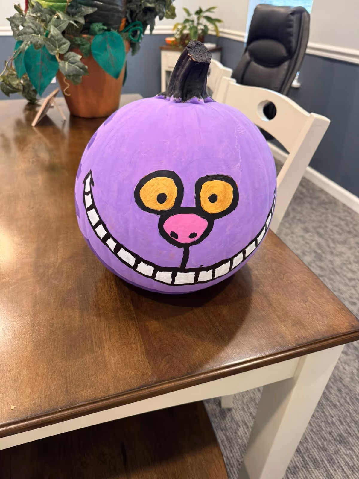 Purple pumpkin with a cartoon face sits on a wooden desk indoors.