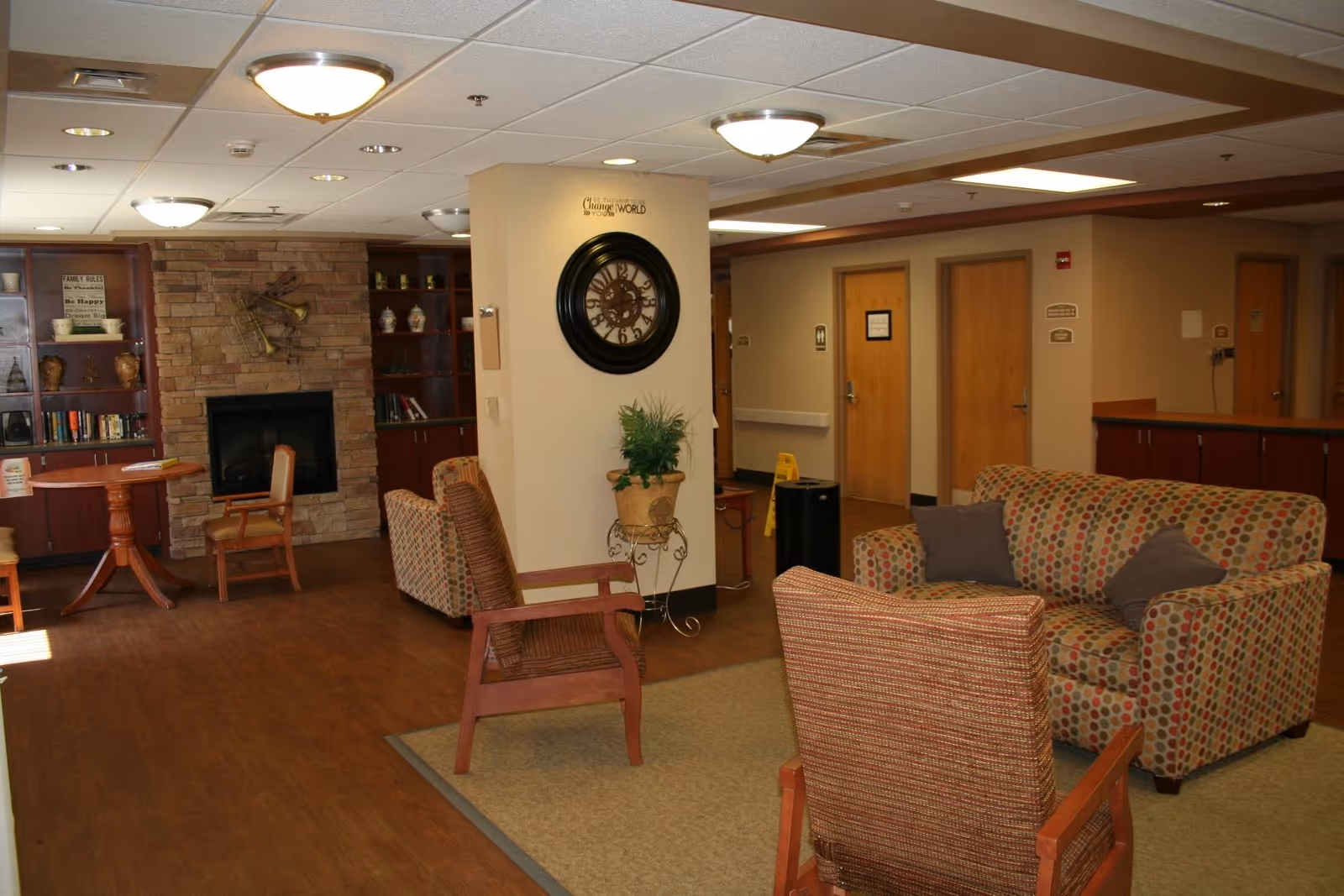 A senior living common room with patterned sofas and chairs, a stone fireplace with bookshelves, and hallway doors.