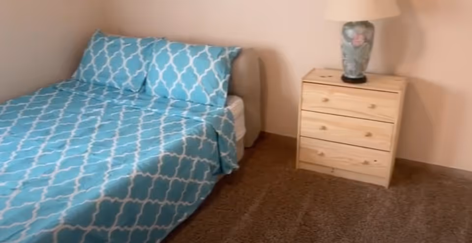 A small bedroom with a blue-patterned bedspread, wooden nightstand, and lamp.