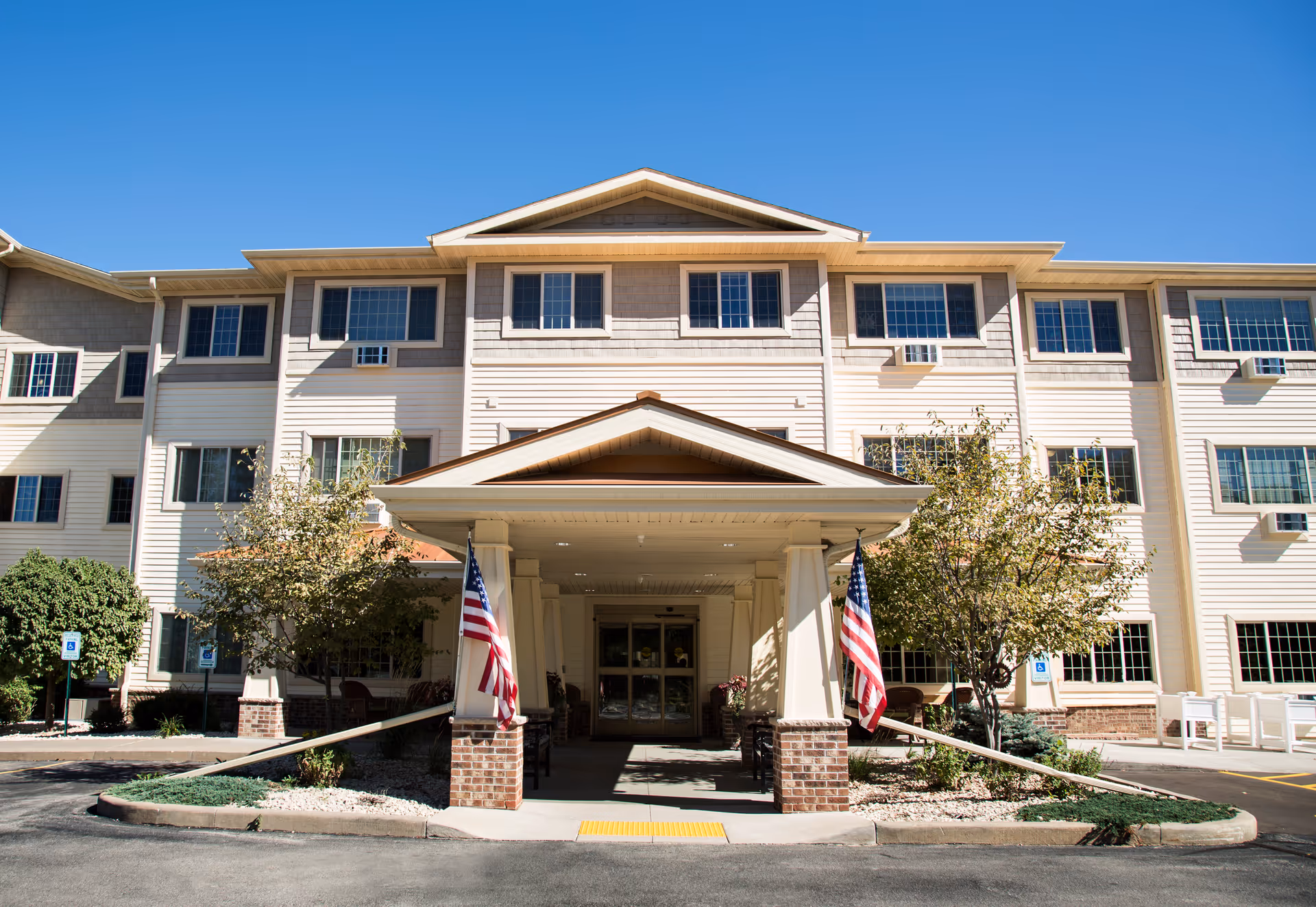 Front exterior view of a three-story assisted living facility building with a covered entrance supported by columns. Two American flags are displayed on either side of the entrance. There are trees and shrubs around the entrance area, and the sky is clear and blue.