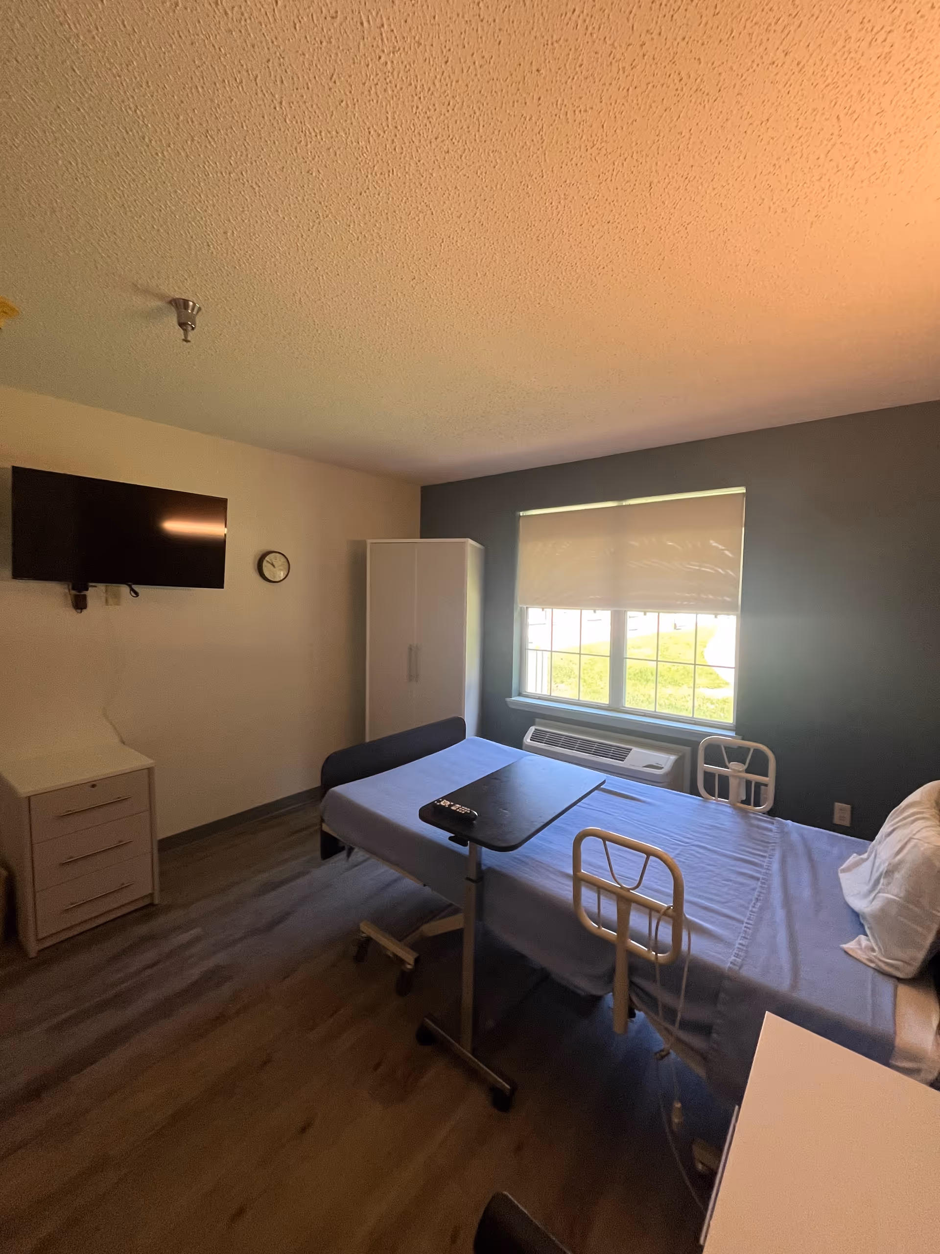 Hospital-style patient room featuring a bed, desk, TV, window, and wall cabinet.