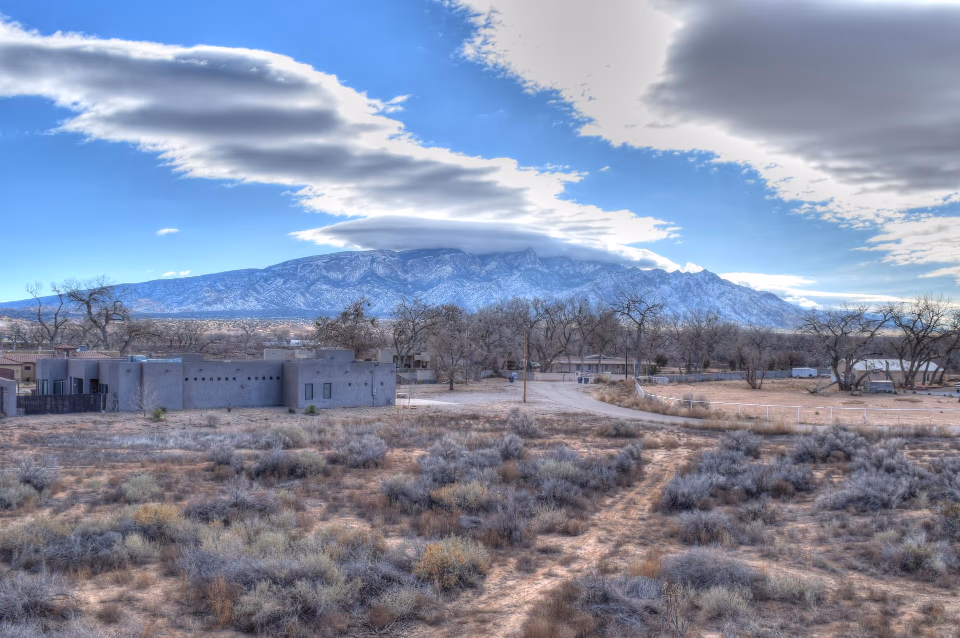 A scenic view of a desert landscape with dry shrubs and a dirt path leading to a cluster of southwestern-style buildings. In the background, there is a large mountain range partially covered with snow under a blue sky with scattered clouds.