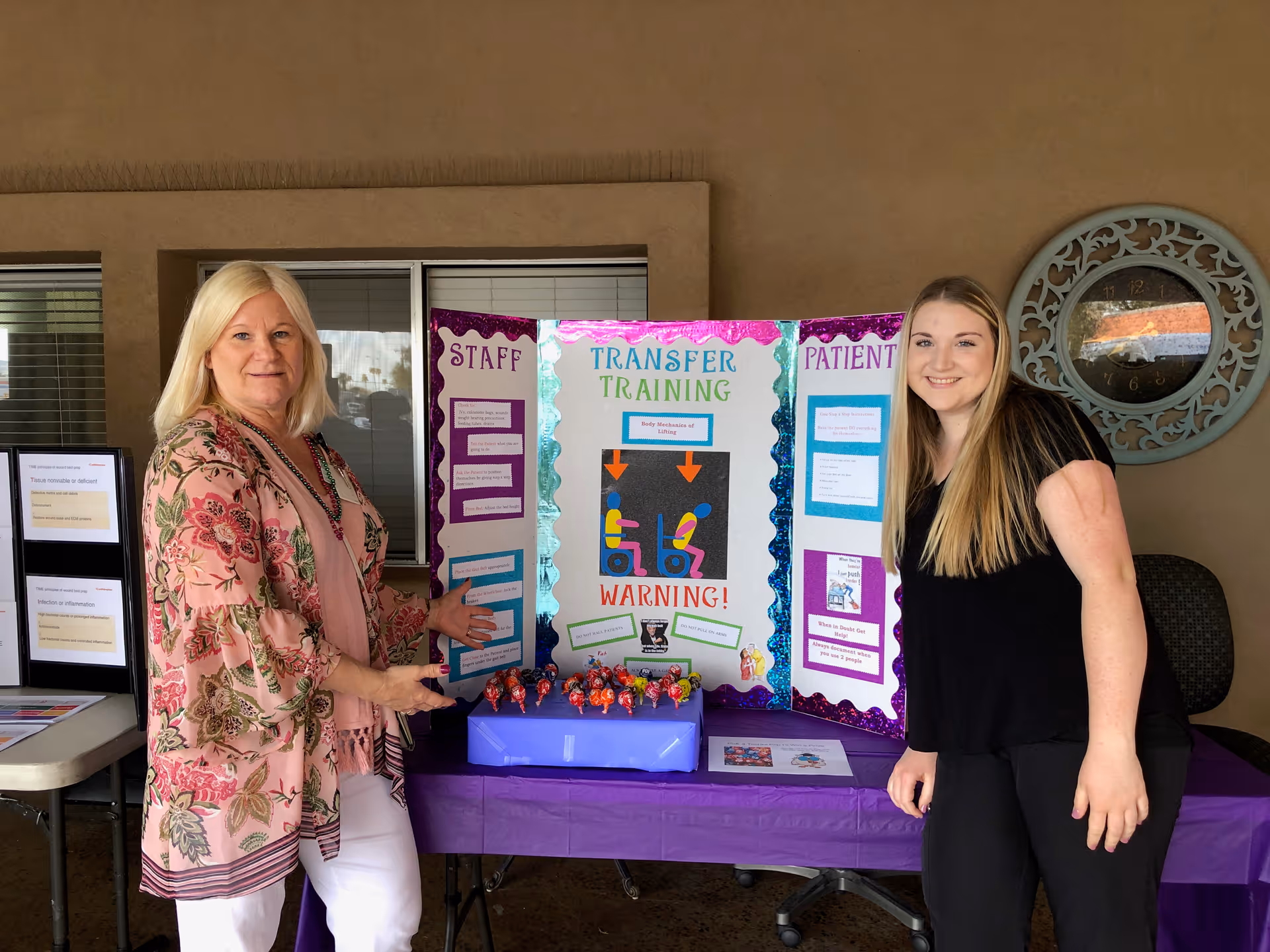 Two women standing on either side of a table with a colorful tri-fold presentation board titled 'Transfer Training' with sections labeled 'Staff' and 'Patient'. The table is covered with a purple tablecloth and has a box of lollipops on it. The setting appears to be indoors with a window and a decorative clock on the wall behind them.