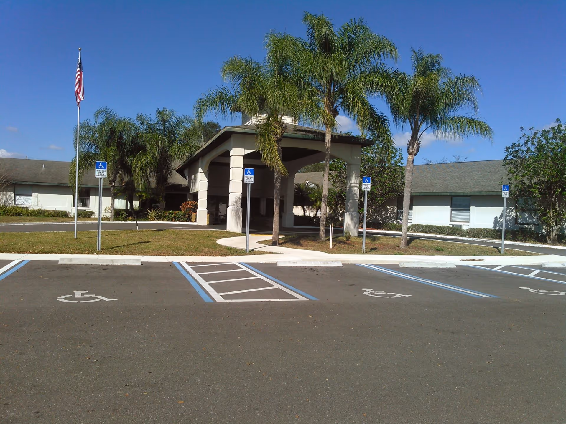 Exterior view of Sunny Hills Alf - Assisted Living Facility Sebring showing a covered entrance with palm trees and several handicap parking spaces in front under a clear blue sky.