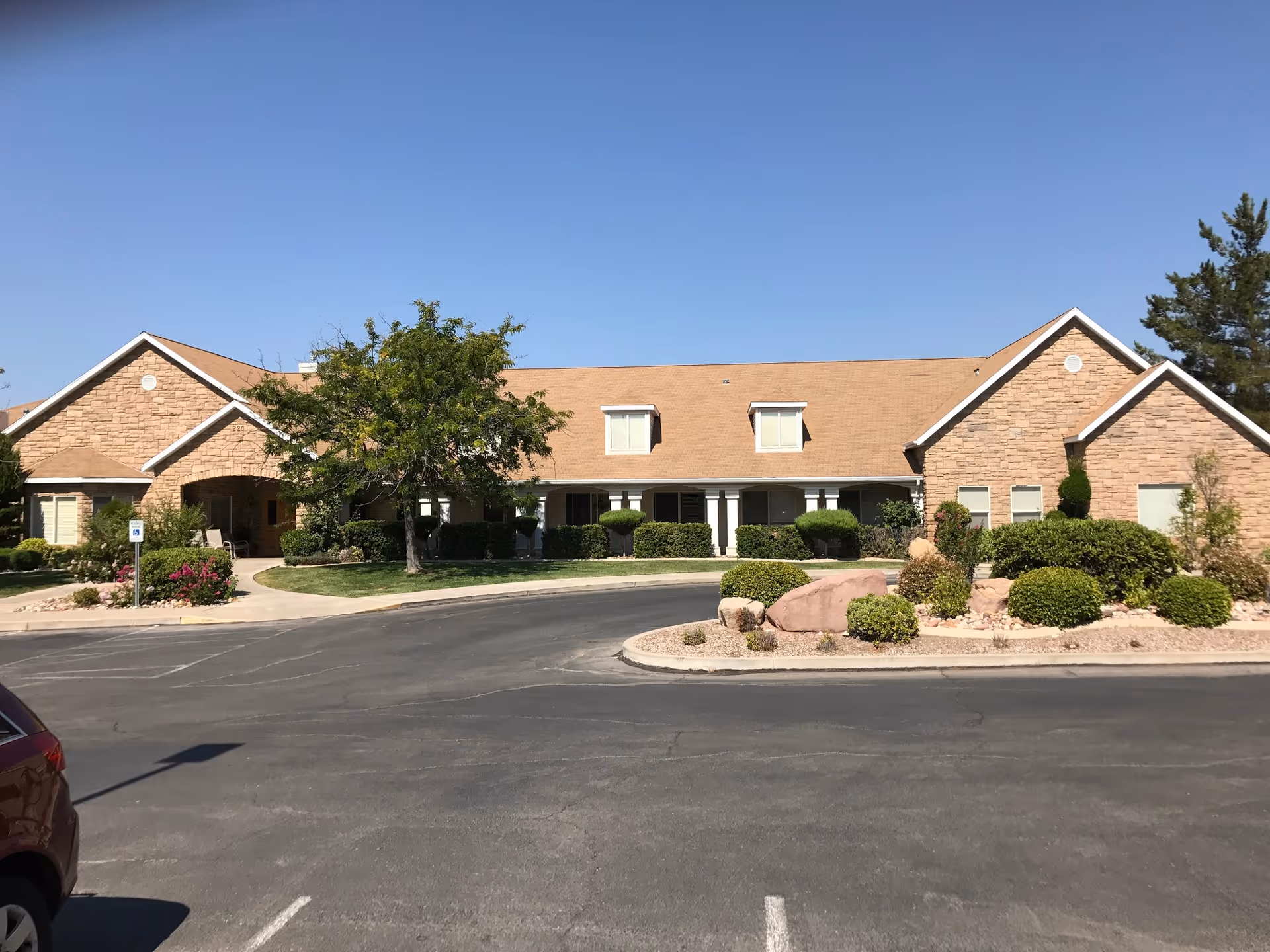 Single-story stone-clad assisted living building front with landscaped shrubs, a driveway, and a clear blue sky.