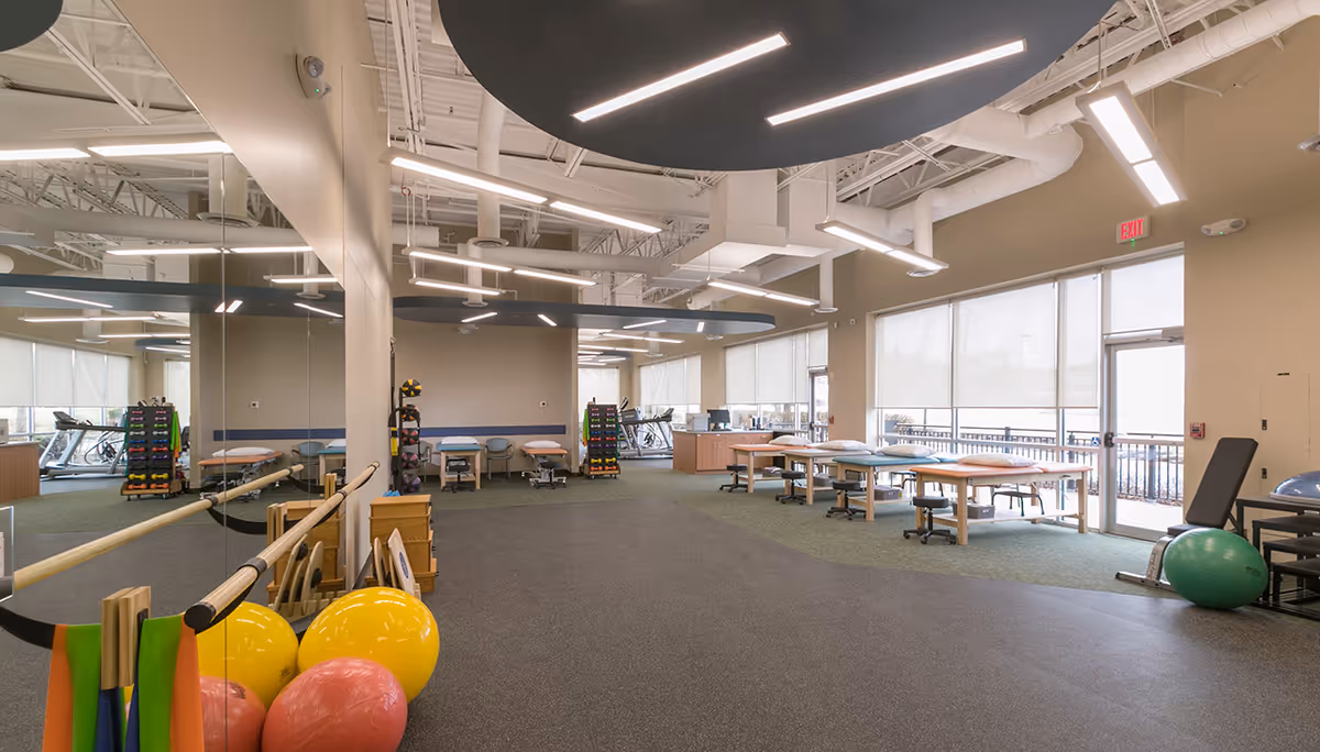 A spacious modern therapy gym with exercise equipment, tables, and bright overhead lighting.