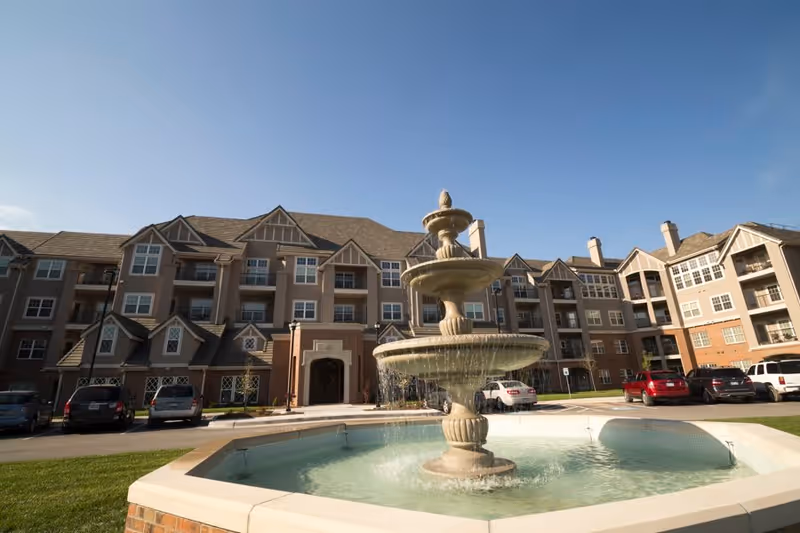 Three-story residential building with a multi-tiered fountain in the foreground, parked cars, and a clear blue sky.