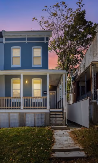 Blue two-story house with front porch, walkway, and tree at dusk.