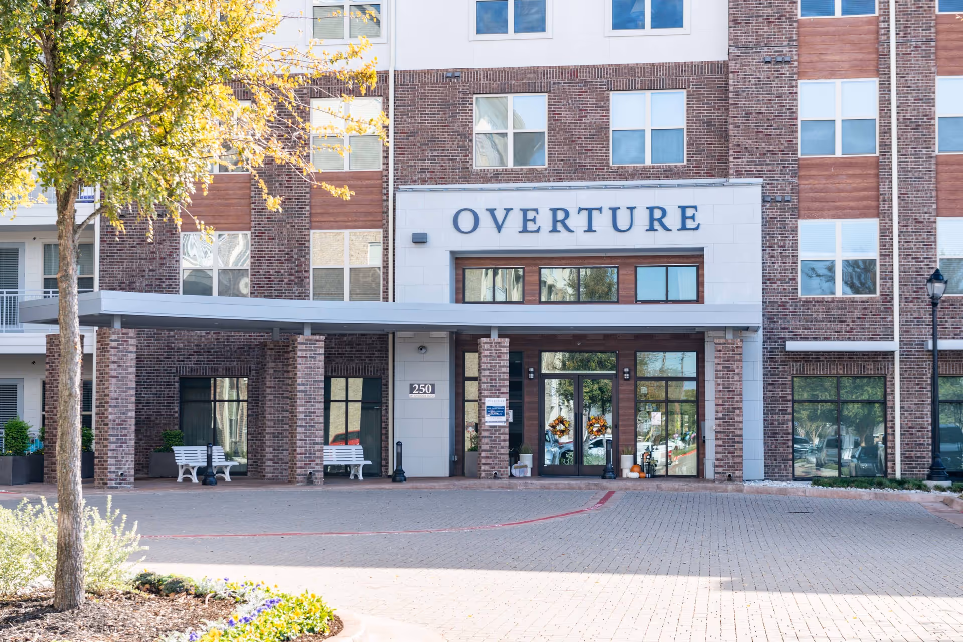 Front entrance of the OVERTURE senior living building with a covered drive, benches, and glass entry doors.