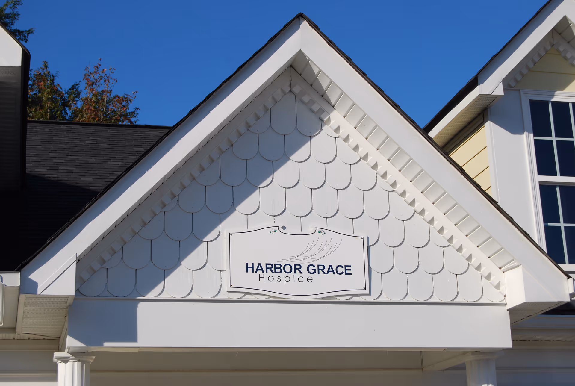White facility front with Harbor Grace Hospice sign on a gabled building under a blue sky.