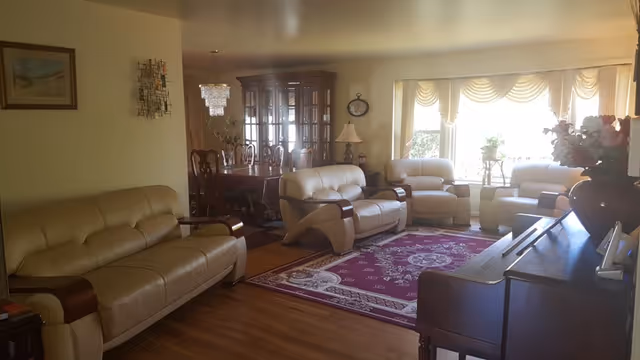 Spacious living room in a senior facility with beige leather seating, a piano, and burgundy rug.