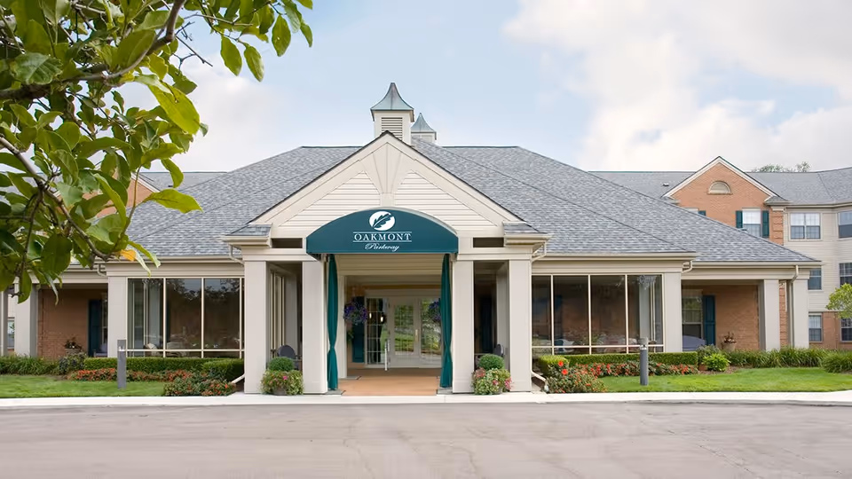 Front exterior view of Oakmont Parkway Senior Community building with a covered entrance, large windows, and landscaped greenery including bushes and flowers.
