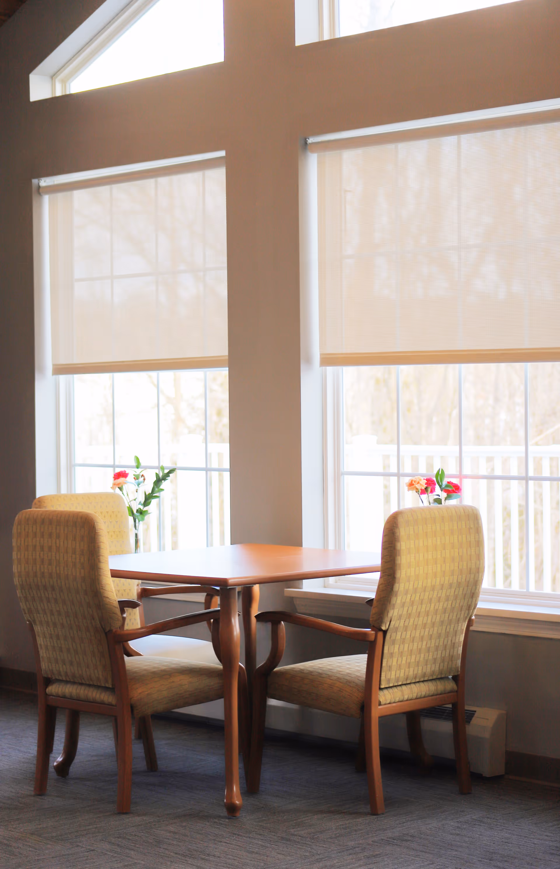 Two upholstered chairs surround a wooden table in a sunlit dining area by large windows.