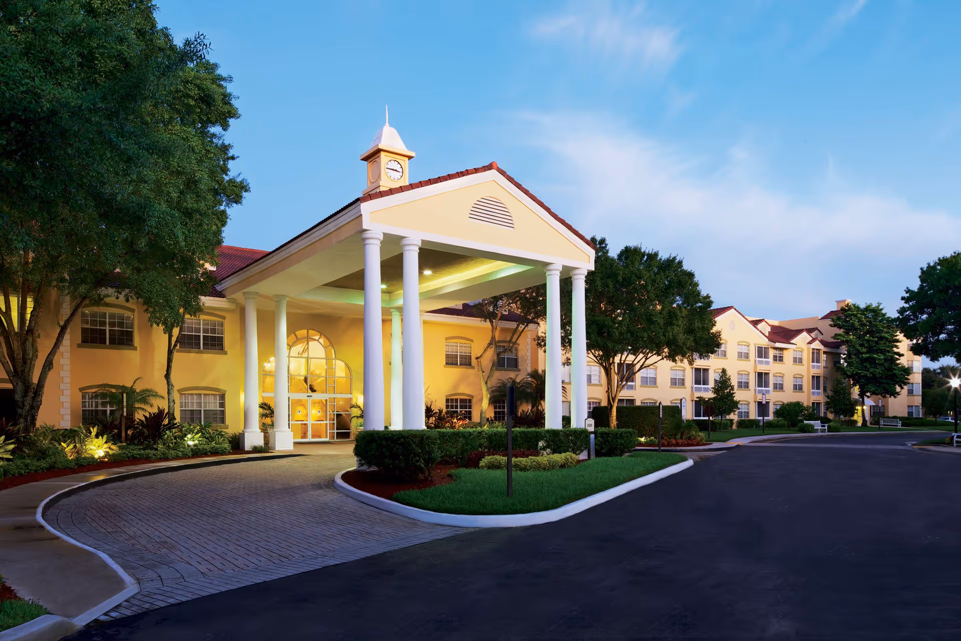 Front entrance of a large yellow residential building with white columns, a covered porte-cochère, clock tower, and surrounding landscaping.