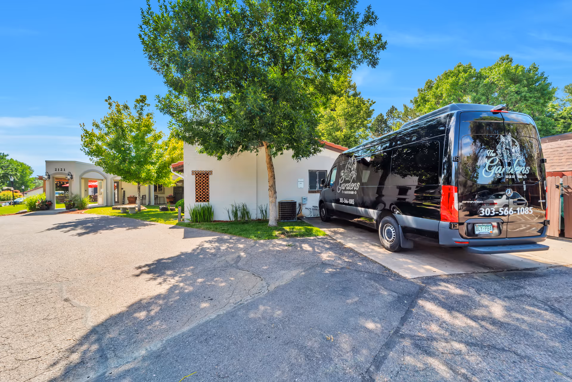 Front exterior of Gardens Care Senior Living showing the entrance, trees, and a black facility shuttle van parked in the driveway.