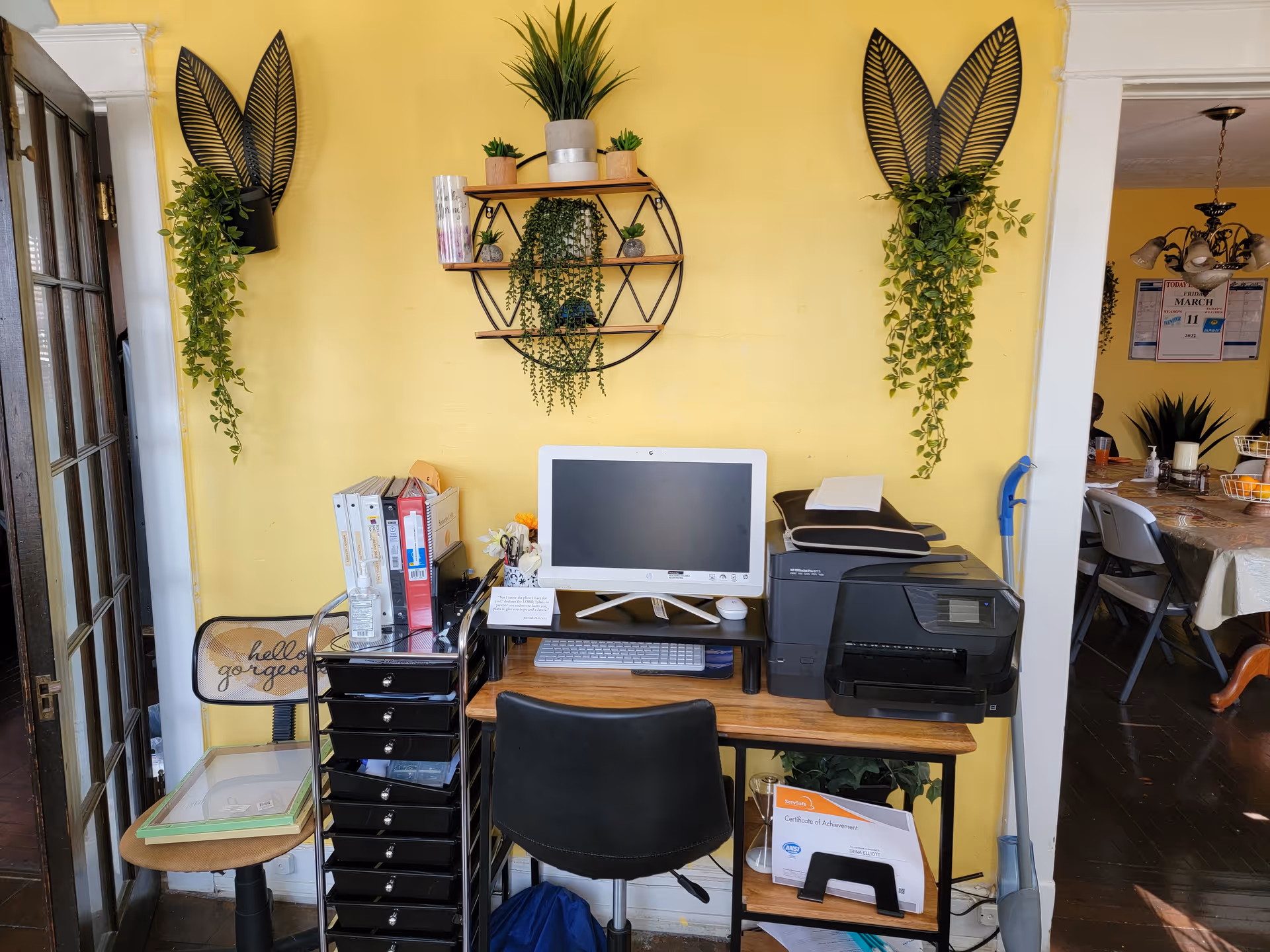 A small office workspace against a yellow wall featuring a computer monitor, keyboard, printer, and several office supplies. The wall is decorated with two black leaf-shaped wall hangings with trailing green plants and a circular shelf holding small potted plants. To the left is a chair with a cushion that says 'hello gorgeous' and a small table with a glass top. To the right, a mop leans against the wall and a dining area with a table and chairs is partially visible.