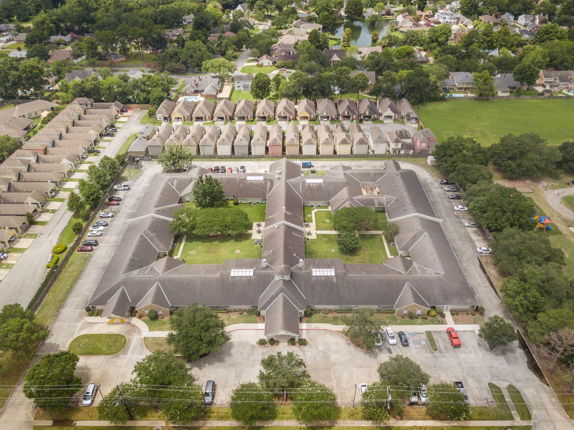 Aerial view of a senior living facility named Brookdale Lafayette, showing a large, U-shaped building with a central courtyard featuring green lawns and trees. Surrounding the main building are parking lots with several cars, rows of smaller residential units, and a neighborhood with houses and greenery in the background.