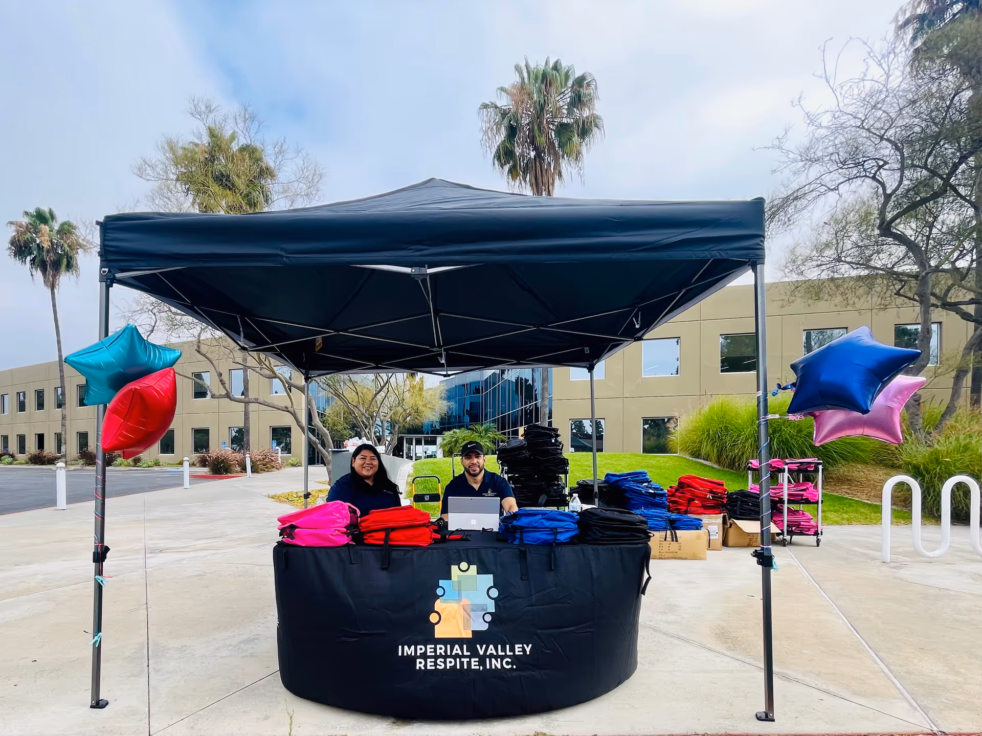 Outdoor setup under a black canopy with staff at a table of colorful blankets and balloons.