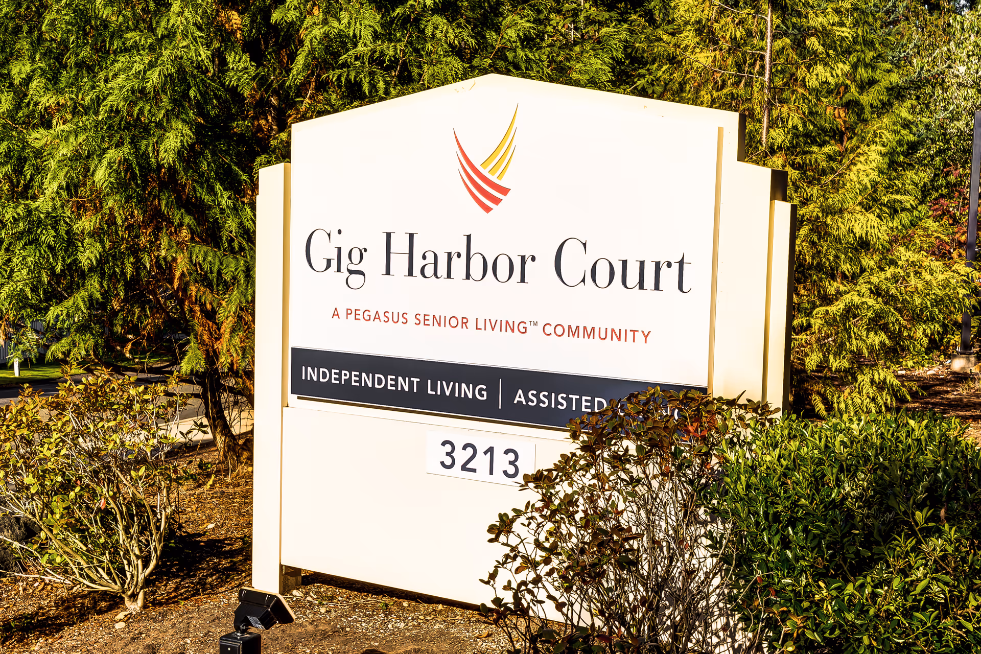 Large white exterior sign reads Gig Harbor Court, a Pegasus Senior Living community, surrounded by greenery.