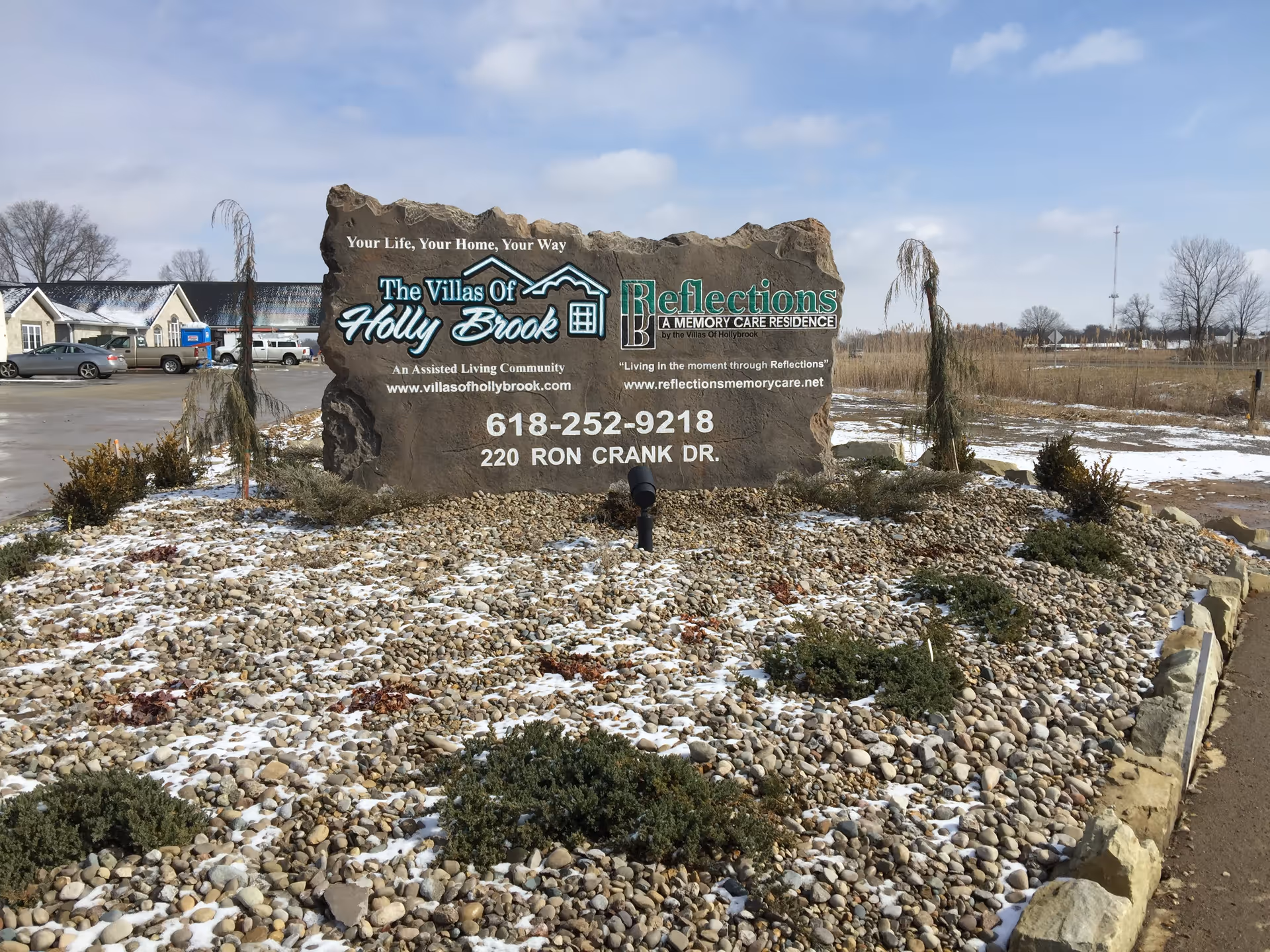 Stone entrance sign for The Villas of Holly Brook and Reflections Memory Care on a rocky landscaped island with light snow.