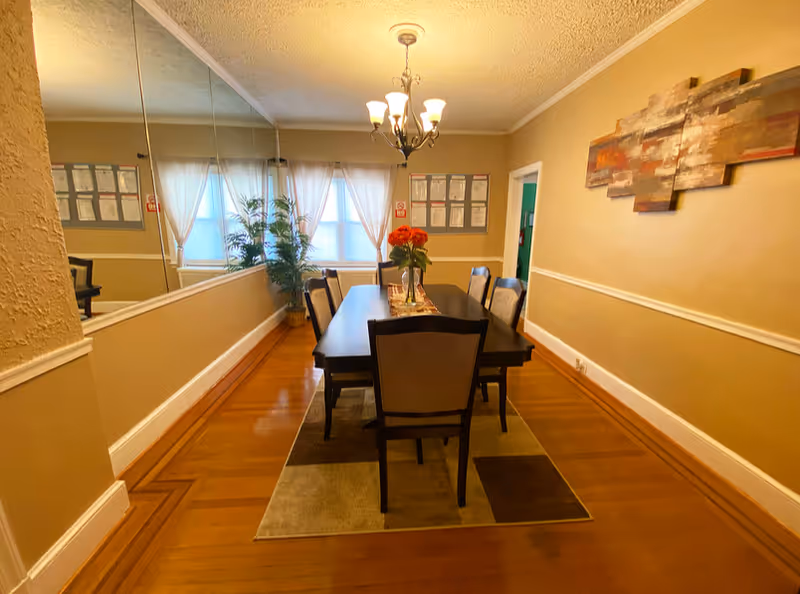 Dining room in a senior living facility with a long table, chairs, chandelier, and plants.