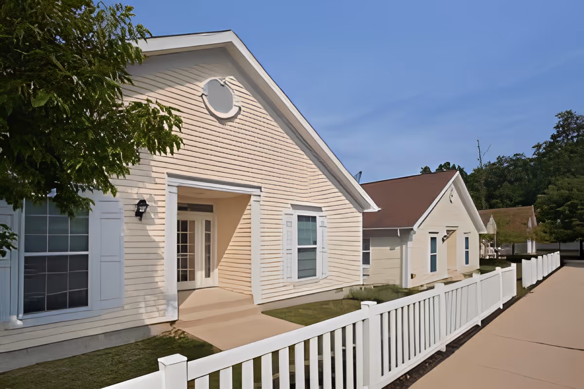 Exterior view of a senior living facility building with beige siding, white trim, and a white picket fence along a paved walkway. The building has a peaked roof and several windows, with trees and greenery in the background under a clear blue sky.