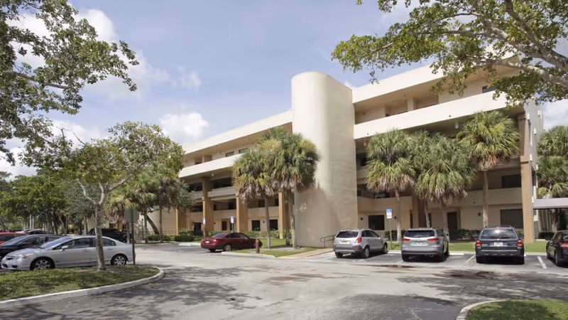 Exterior view of a multi-story senior care building with palm trees and parked cars.