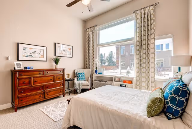 A bright and cozy bedroom with a large window letting in natural light. The room features a wooden dresser with framed pictures and a plant on top, a comfortable armchair with a blue patterned pillow, and a neatly made bed with white bedding and decorative pillows in blue and green. Light-colored curtains frame the window, and a ceiling fan is visible above.