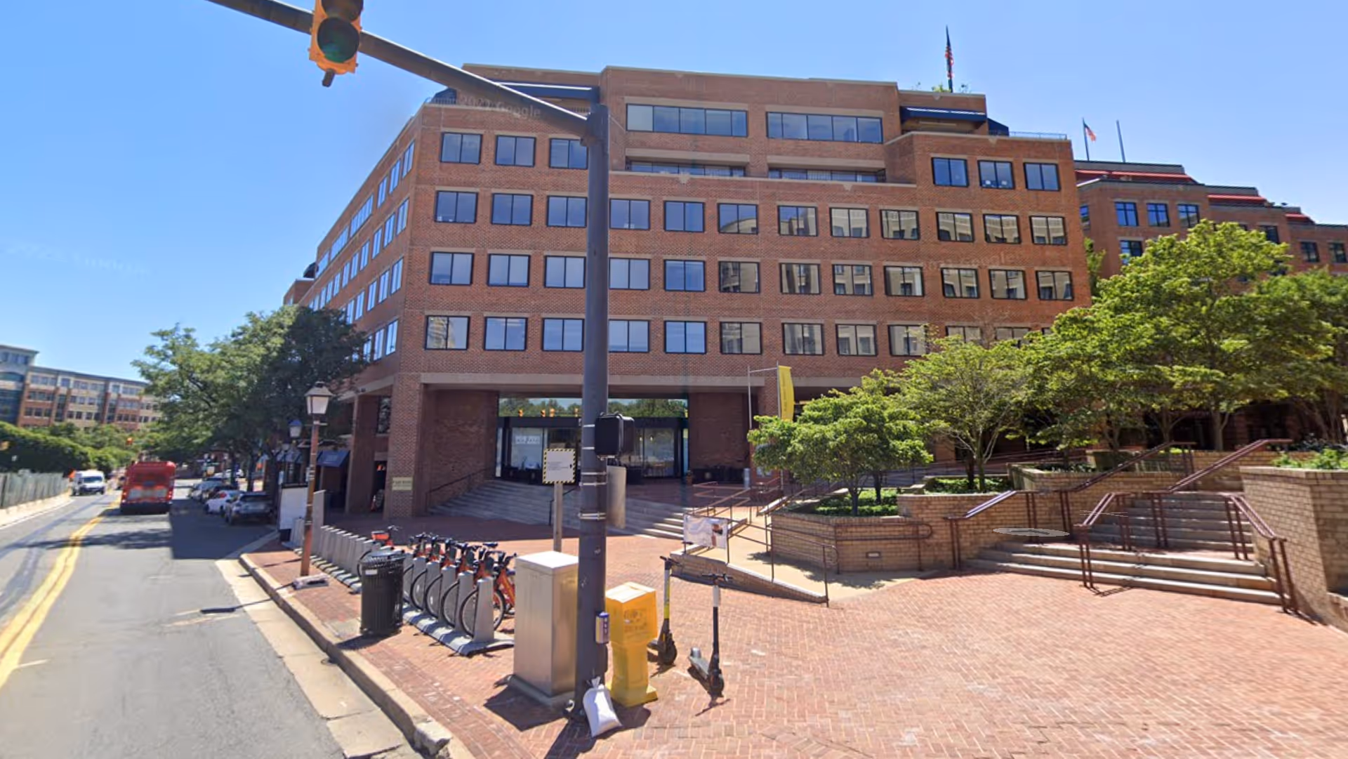 Brick, multi-story building facade with steps, trees, and a brick plaza beside a city street.