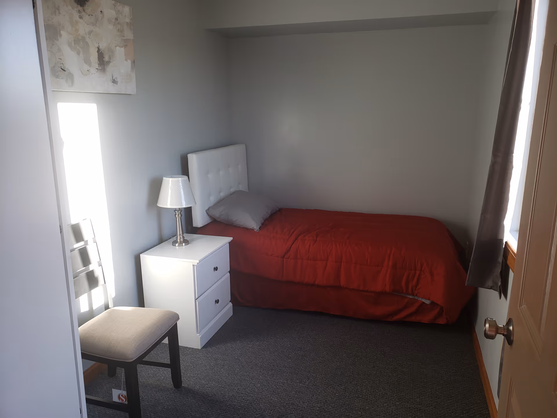 Small bedroom in senior living facility with a single bed, red comforter, nightstand, lamp, and chair