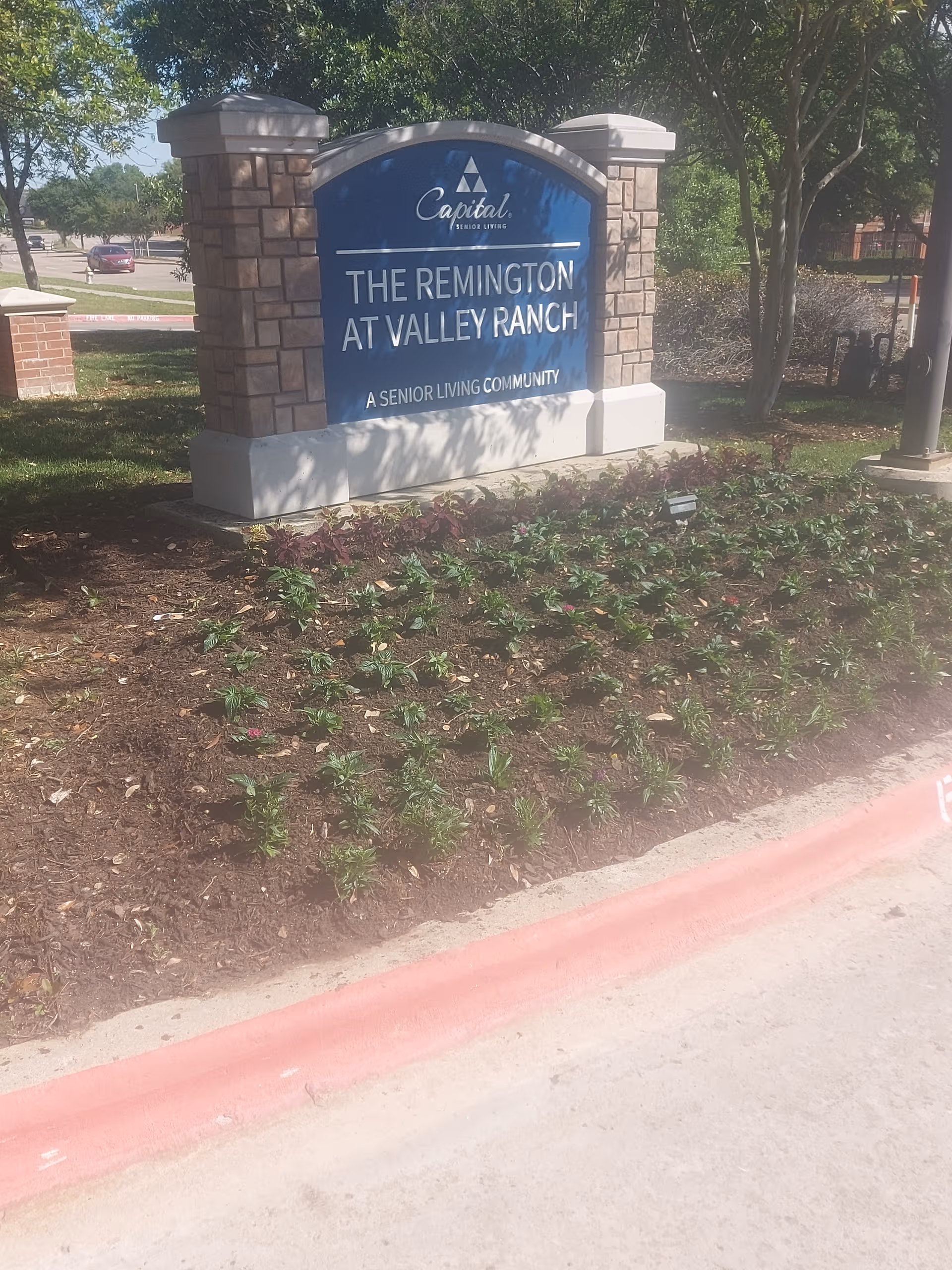 Blue sign reads The Remington at Valley Ranch, set among landscaped shrubs near the curb.