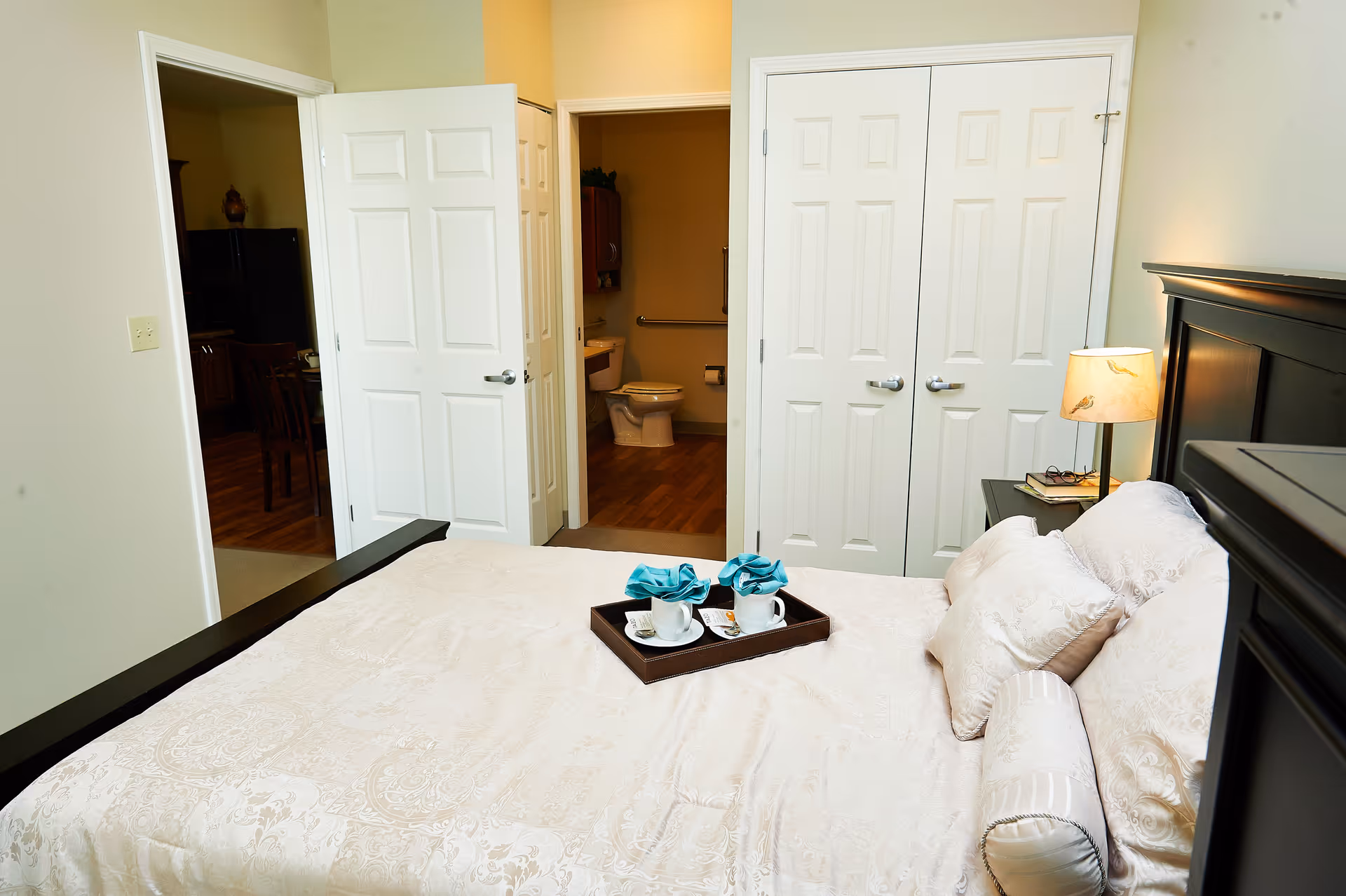 A bedroom in a senior living facility with a neatly made bed featuring cream-colored bedding and decorative pillows. A tray with two cups and napkins is placed on the bed. The room has a bedside table with a lamp and books. In the background, there are two open doors, one leading to a bathroom with a visible toilet and sink, and the other leading to another room with wooden flooring and furniture.