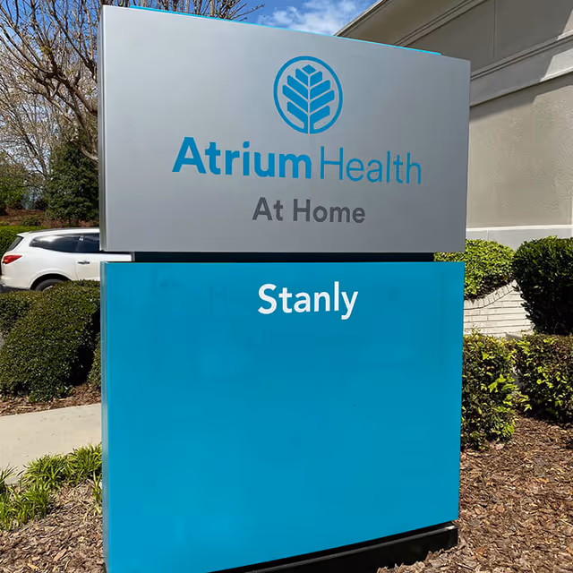 Outdoor sign for Atrium Health At Home Stanly beside a building and landscaping.